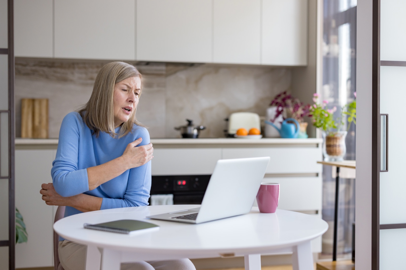Woman holding her shoulder while using a laptop for an online frozen shoulder treatment consultation